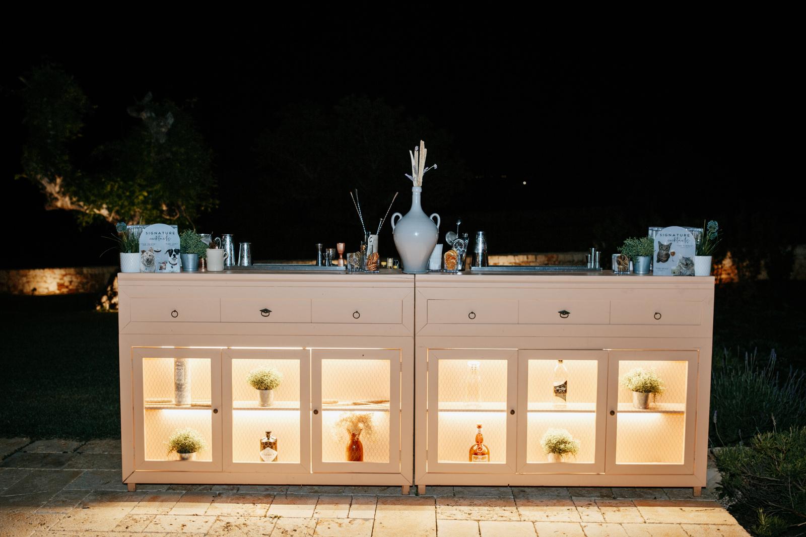 terracotta station evento elegante Bancone bar Terracotta Station by JoeBar, allestito di sera all’aperto con bottiglie illuminate, piante aromatiche e dettagli in ceramica pugliese.